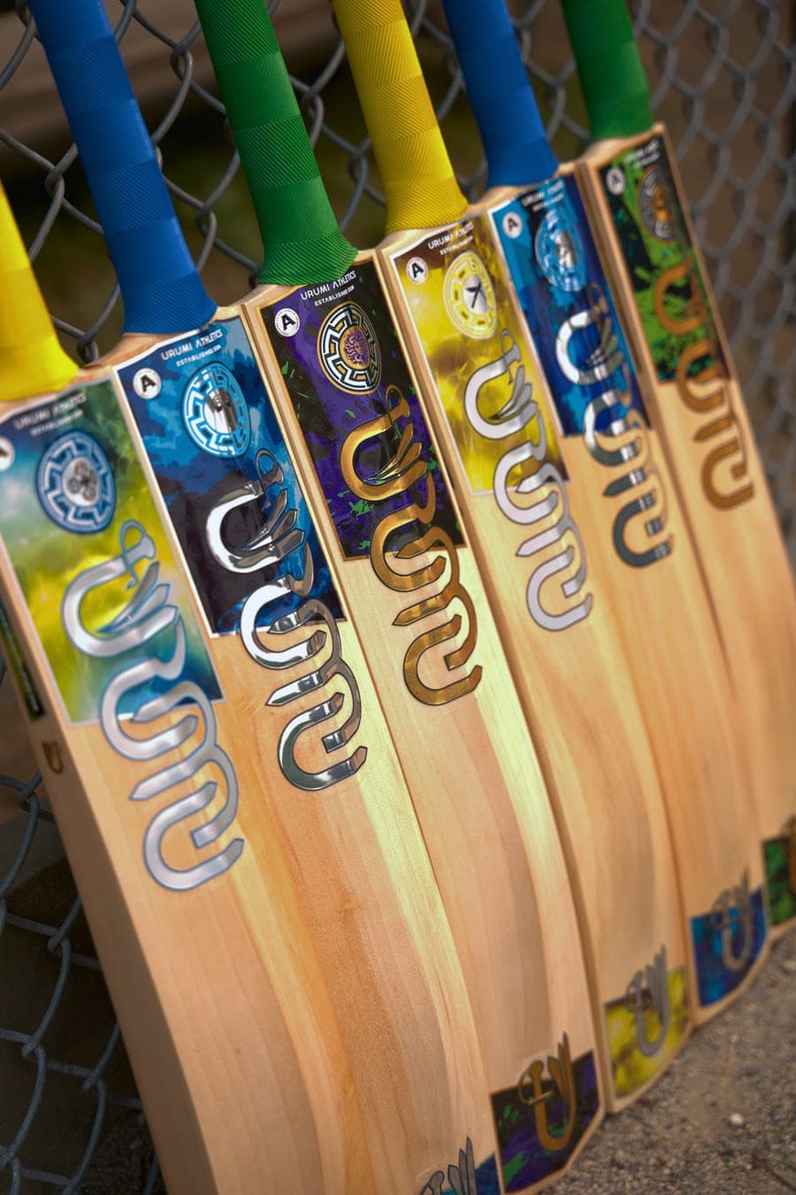 Urumi cricket bats with blue, green and yellow grips lined up at the nets